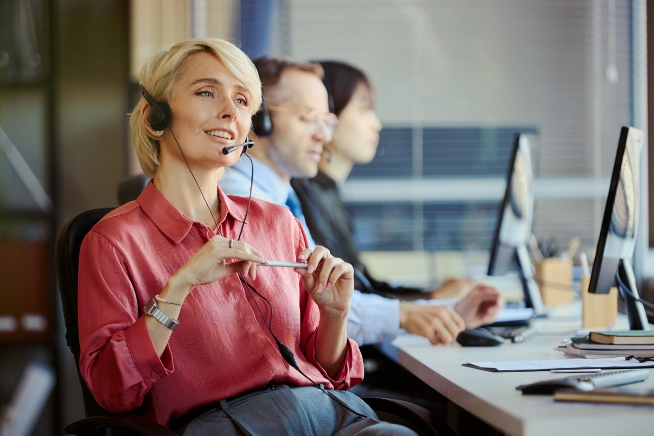 A worker wearing a telephone headset at a desk, colleagues at workstations behind her
